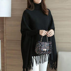 Person wearing a black poncho holding a patterned handbag against a wooden wall.
