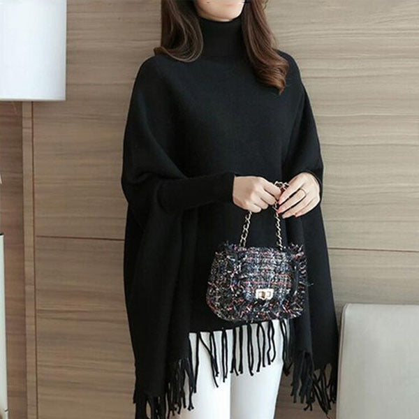 Person wearing a black poncho holding a patterned handbag against a wooden wall.