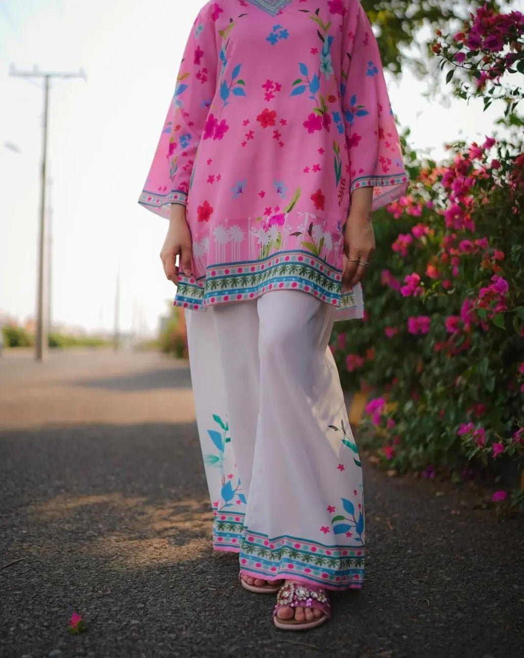 Person wearing a pink floral top and white pants with floral patterns, standing outdoors.