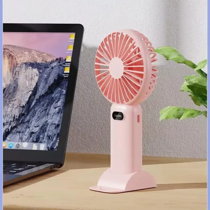 Pink handheld fan on a desk next to a laptop with a mountainous landscape on the screen.