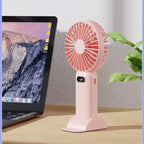 Pink handheld fan on a desk next to a laptop with a mountainous landscape on the screen.