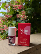 Benefit bene tint lip and cheek stain with packaging on a wooden surface with flowers in the background