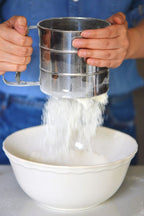 Person sifting flour into a white bowl using a metal sifter.