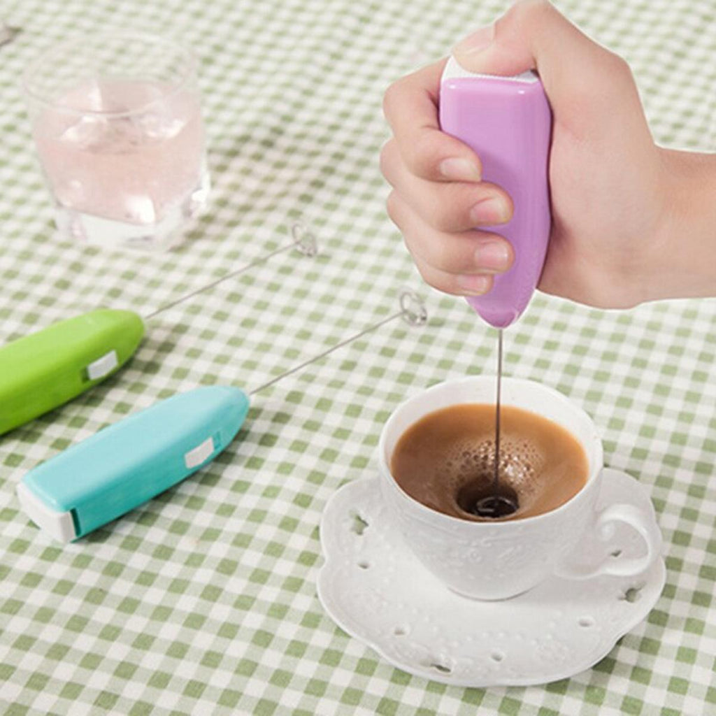 Hand using a purple milk frother to create foam in a cup of coffee on a green checkered tablecloth.