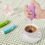 Hand using a purple milk frother to create foam in a cup of coffee on a green checkered tablecloth.