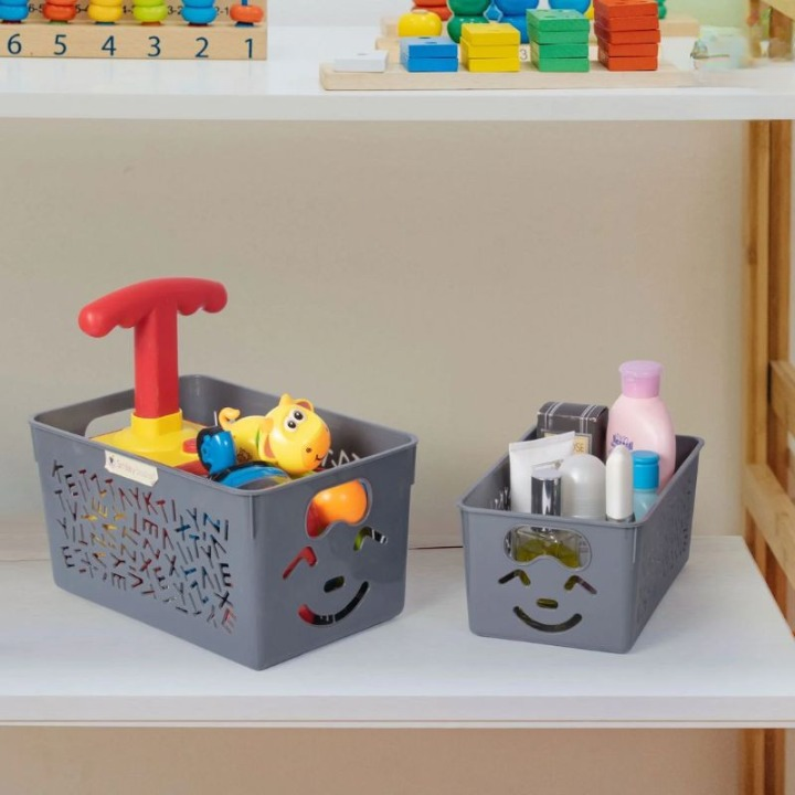 Two gray storage bins with smiley face designs, one containing toys and the other containing bottles, on a white surface.