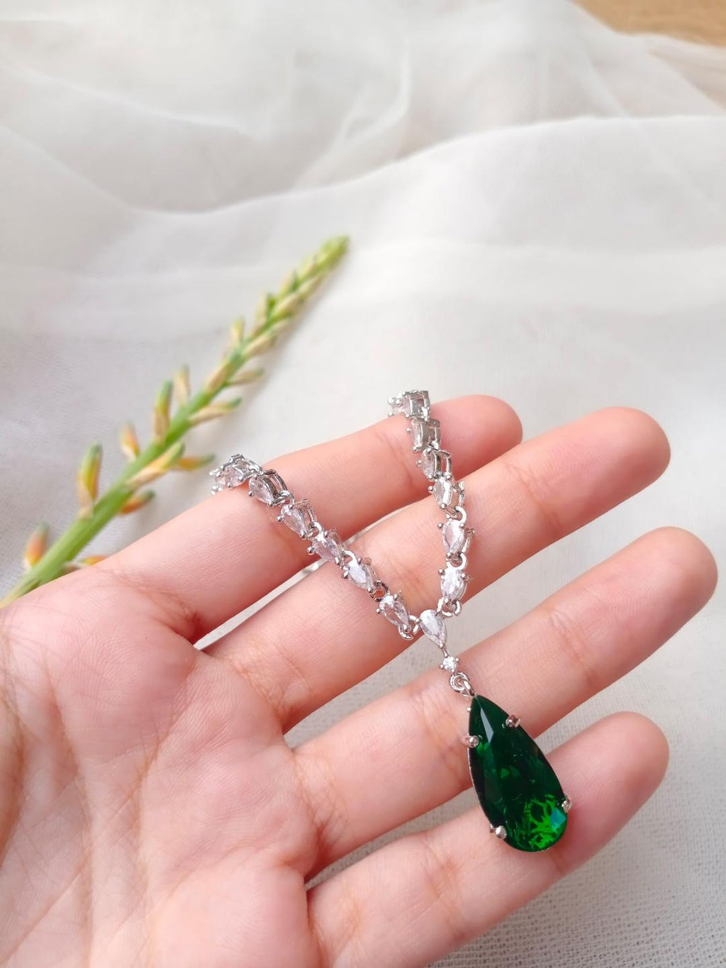 Hand holding a silver necklace with a green teardrop pendant on a light fabric background