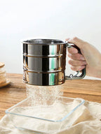Hand holding a metal sifter over a glass container on a wooden surface.