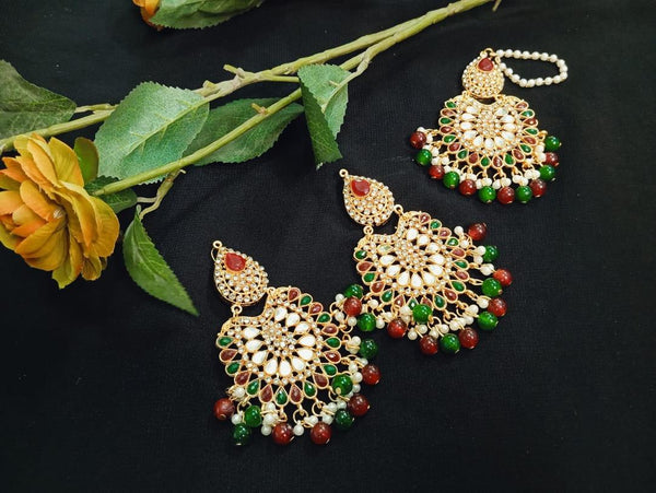 Ornate gold earrings with green and red stones on a dark surface with a yellow flower and green leaves.